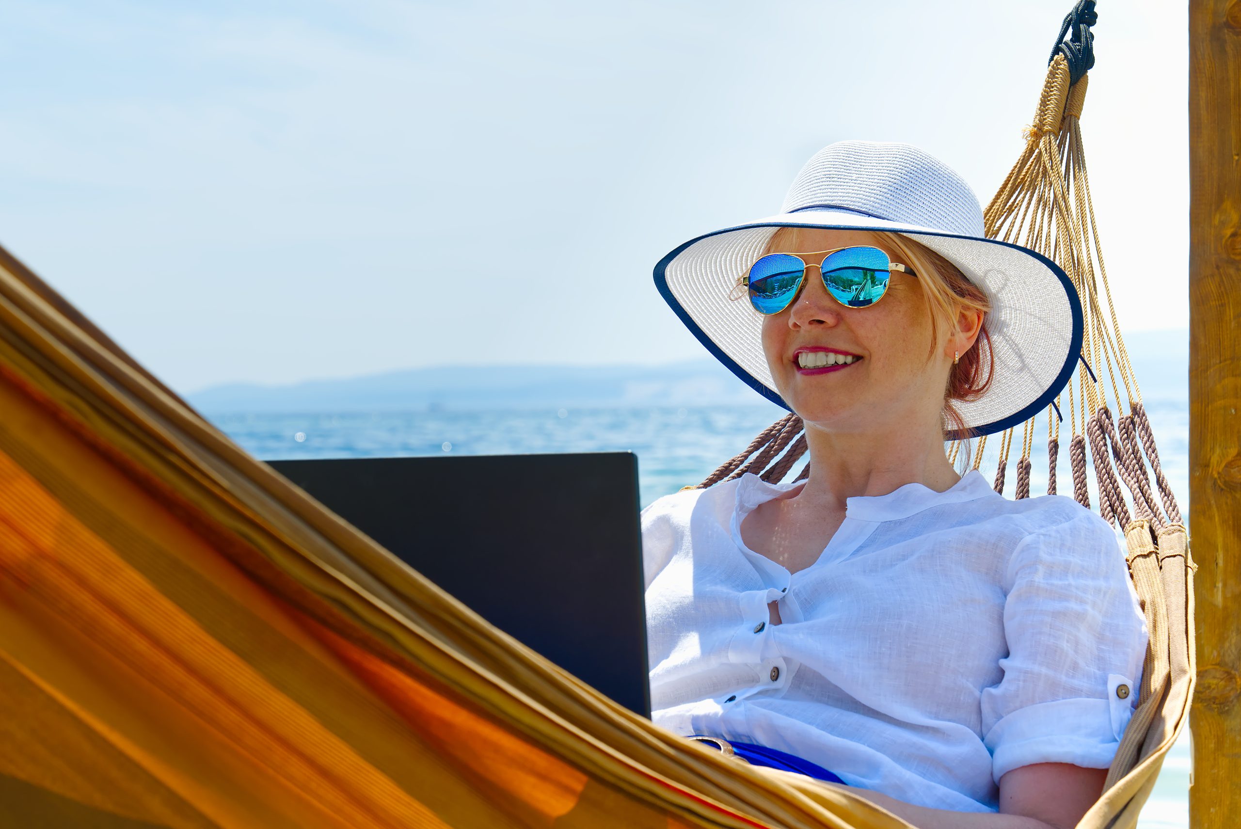 Work from anywhere. Young smilyng woman, female freelancer in straw hat working on laptop on beach