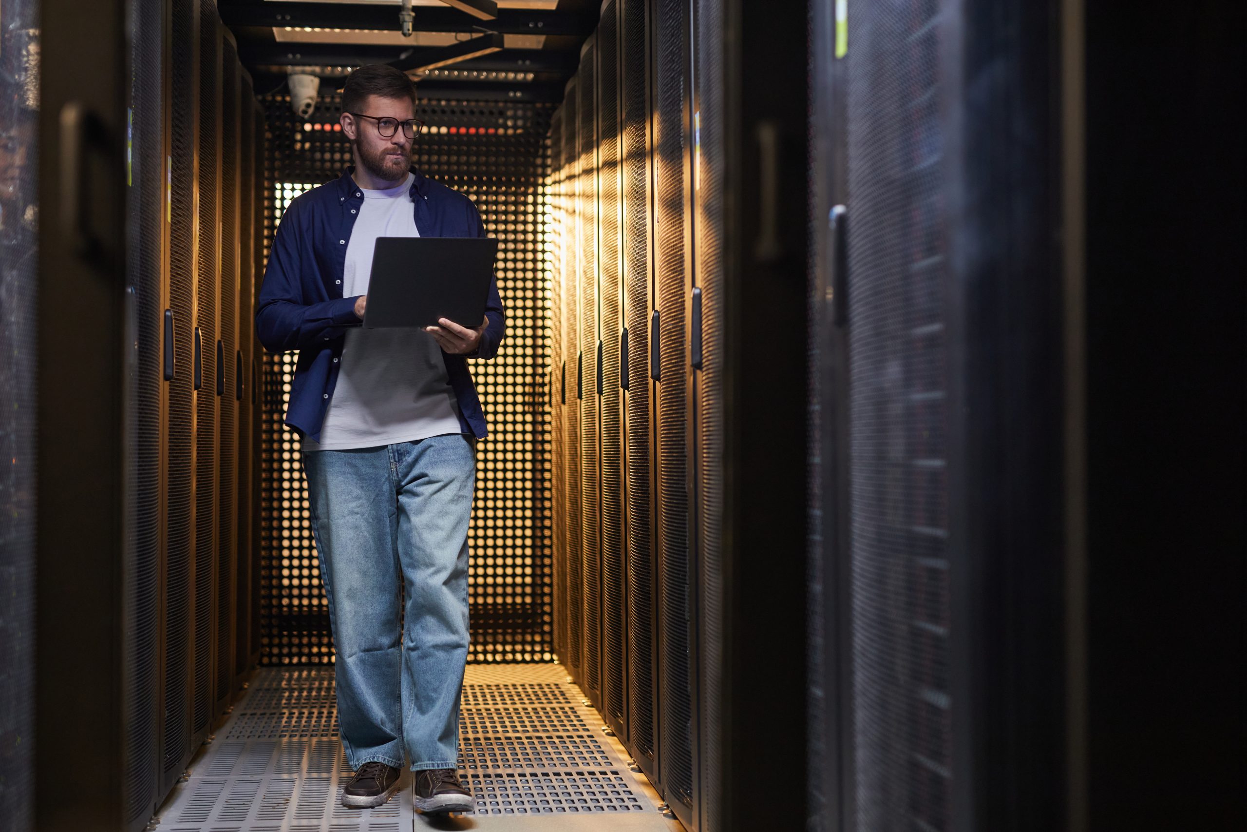 Technician standing in server aisle while monitoring data systems on laptop. Surrounded by server racks, ensuring smooth operation of network systems